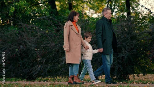 Happy child holding hands with mom dad walks through autumn park. Healthy family walk, mother, child, father in park. Kid son, mom, dad walk together holding hands in autumn outdoor. Mother father boy
