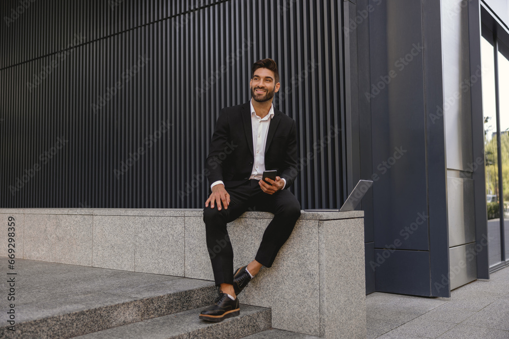 Businessman with mobile phone sitting on bench on office building background. High quality photo Businessman with mobile phone sitting on bench on office building background. High quality photo