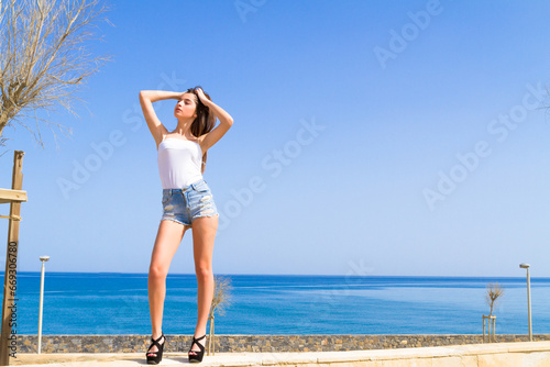 Confident stylish beautiful brunete young girl full length photo, wearing trendy casual shredded blue jeans shorts, white top and black elevated shoes, posing at urban park in blue ocean background.