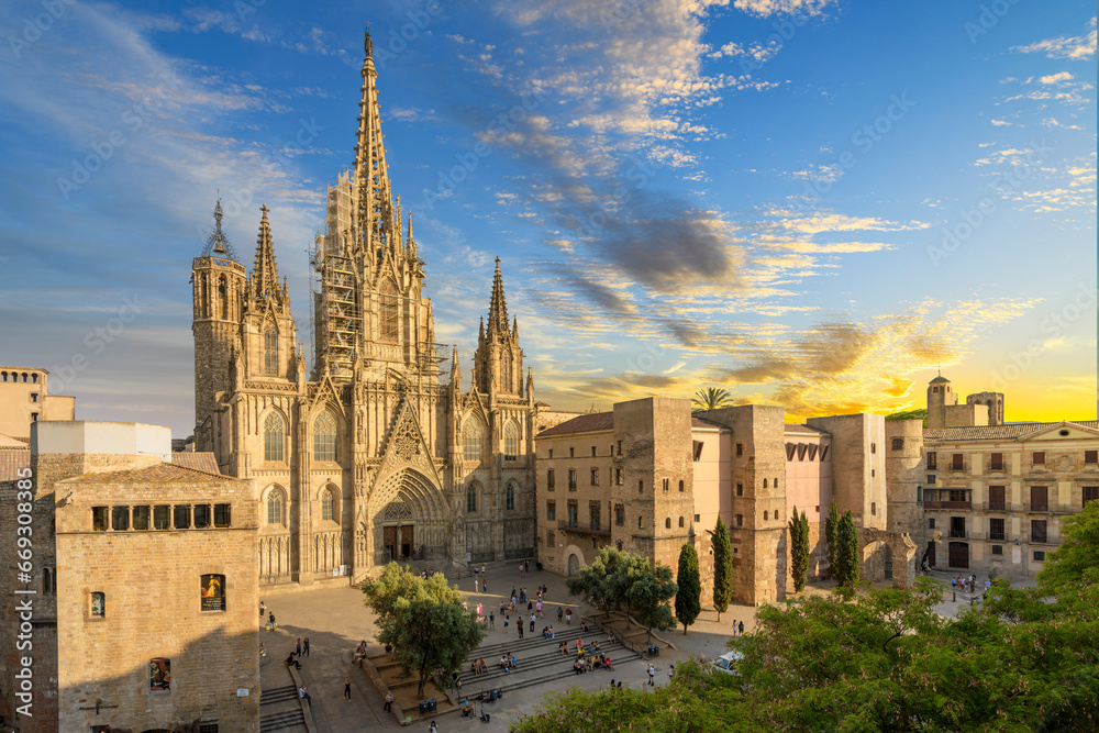 View of the Gothic Barcelona Cathedral of the Holy Cross and Saint Eulalia with surround buildings, plaza and the skyline of Barcelona in view as the sun sets at dusk.