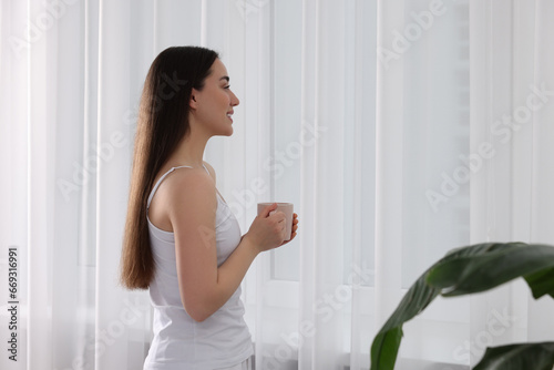 Happy woman with cup of drink near window indoors  space for text. Lazy morning