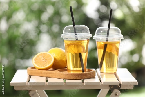 Plastic cups of tasty iced tea with lemon and fresh fruits on white wooden table against blurred green background © New Africa