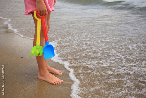 Little girl with plastic toys standing on sandy beach near sea, closeup. Space for text
