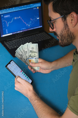 Operador, Trader de Forex. Hombre joven haciendo Trading con laptop, teléfono móvil y dólares sobre superficie azul. Mercado de divisas. Binance, Cripto, Bitcoin.  photo