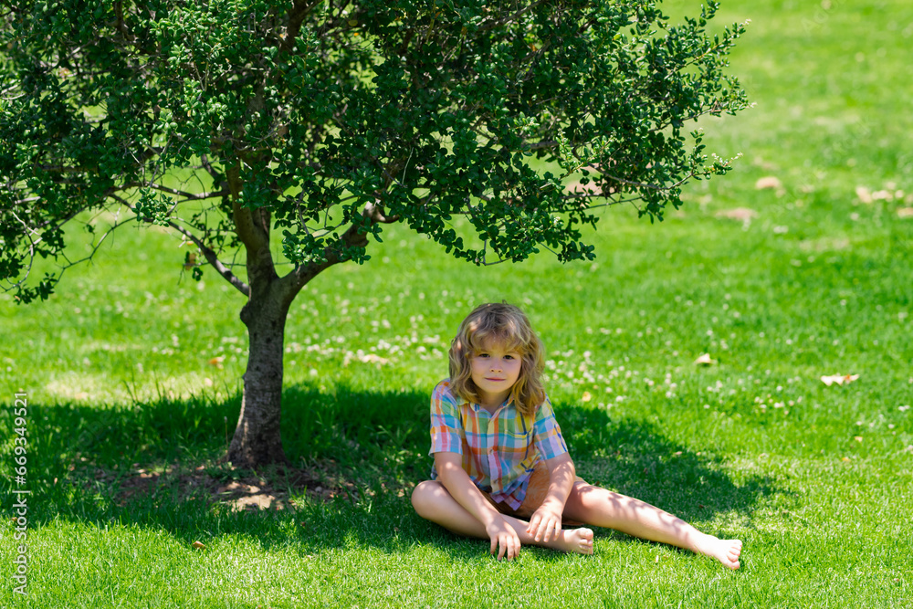 Cute child boy enjoy relax on lawn. Child playing in garden. Happy little boy sitting on the grass at the summer day.