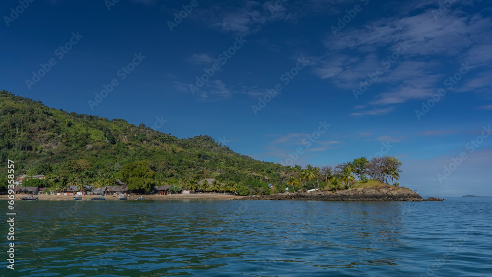 A picturesque tropical island against a background of blue sky and clouds. Lush green vegetation on the hill. Houses with thatched roofs near the shore. The boats are moored at the water's edge. 