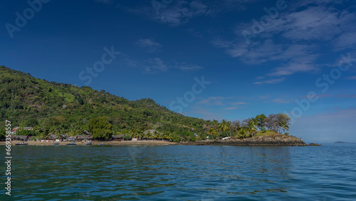 A picturesque tropical island against a background of blue sky and clouds. Lush green vegetation on the hill. Houses with thatched roofs near the shore. The boats are moored at the water's edge. 
