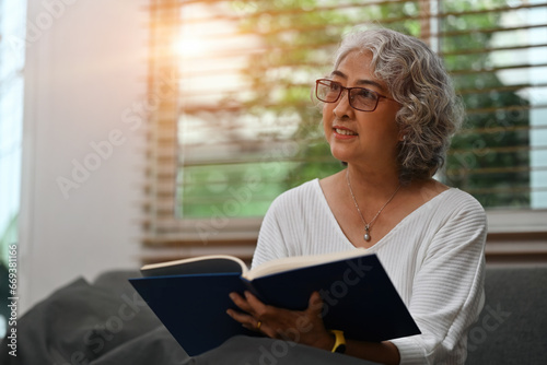 A smiling Senior Asian woman relaxes on the couch in the living room while reading a book, or literature, enjoying free time on the weekend. Elderly people's lifestyles and leisure activity concepts