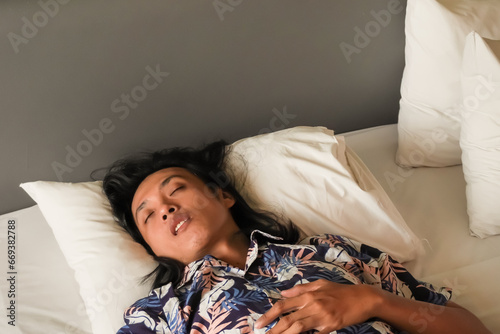 An Asian man with long hair is sleeping on your back with his mouth open on a bed with a white pillow. Sleep comfortably at the hotel during holiday vacation. Rest after a long journey photo