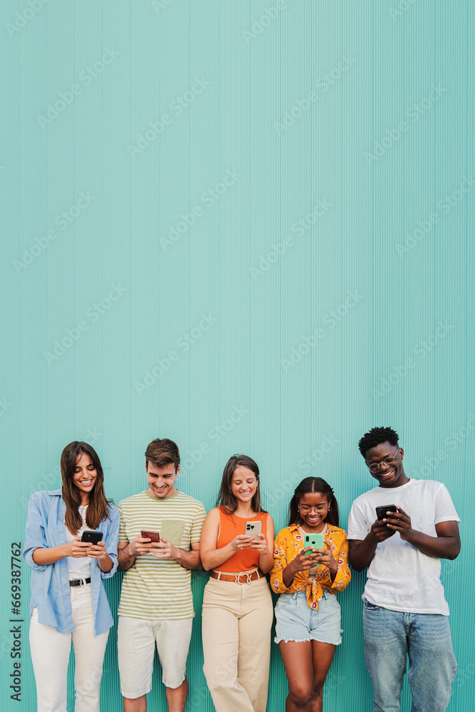 Vertical. Multiracial group of young friends smiling using their mobile phone app at teal wall. Teenagers having fun sharing messages on social media with a cellphone at blue color background. High Vertical. Multiracial group of young friends smiling using their mobile phone app at teal wall. Teenagers having fun sharing messages on social media with a cellphone at blue color background. High