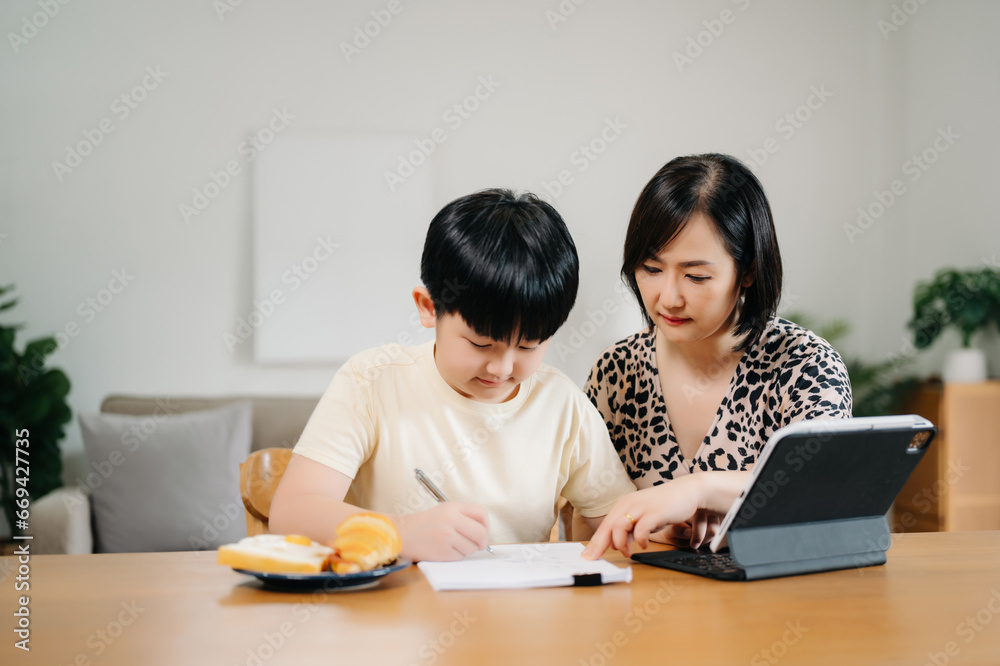 Homeschool Asian young little boy learning online and does homework by using computer and tablet with mother help, teach and encourage. son smile to study at home together with mom Homeschool Asian young little boy learning online and does homework by using computer and tablet with mother help, teach and encourage. son smile to study at home together with mom