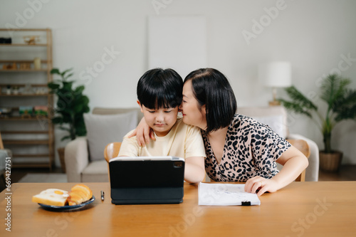 Homeschool Asian young little boy learning online and does homework by using computer and tablet with mother help, teach and encourage. son smile to study at home together with mom