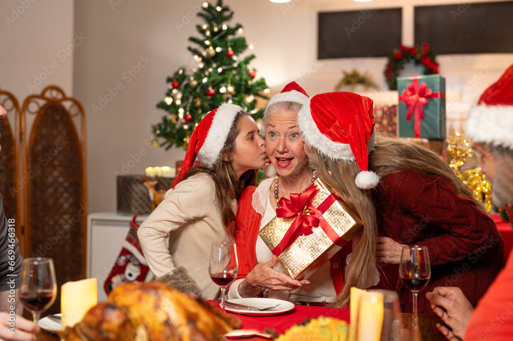 Happy family celebrating Christmas dinner at home. Granddaughter with gift box surprise given to grandmother wearing Santa hats. Merry Christmas or New Year's eve concept. Happy family celebrating Christmas dinner at home. Granddaughter with gift box surprise given to grandmother wearing Santa hats. Merry Christmas or New Year's eve concept.