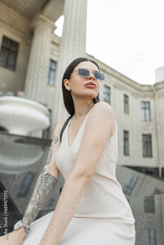 Beautiful stylish urban model woman with vintage gold sunglasses in a fashion beige dress with a tattoo on her arm sits in the city near a retro columns building