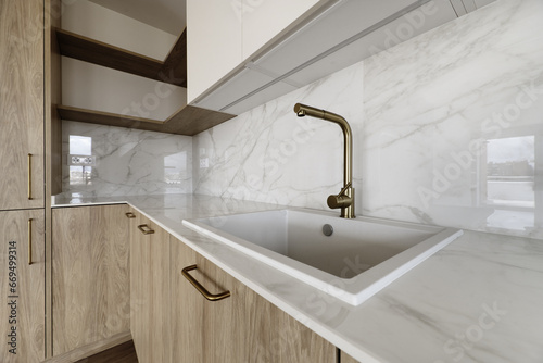 Corner of a newly installed modern kitchen with a white resin sink with a gold metal faucet on a quartz-like stone countertop and light wood cabinets