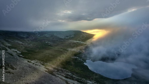Aerial drone shot of sunrise in the mountains, fiery yellow sun rays breaking through the fog and white clouds. Flying over tundra landscape with mountain river and lake, Swedish wilderness