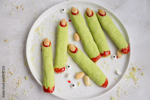 Witches fingers cookies for Halloweem in green color and with red jelly blood on white plate and background close up selective focus