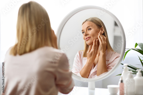 Facelift Massage. Beautiful Middle Aged Woman Massaging Face While Standing Near Mirror photo