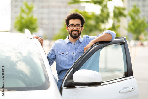 Happy indian guy standing by white car at parking, smiling