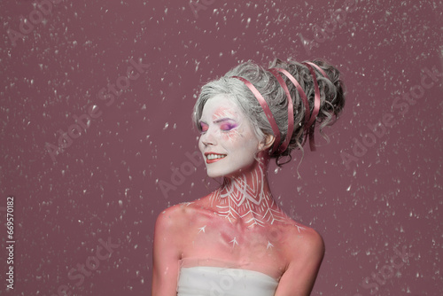 Happy winter woman actress with stage makeup, pink painted body and perfect hairstyle having fun under snow fall on pink background, portrait photo