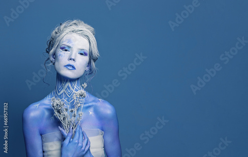 Glamorous winter queen woman actress with stage holding dried flower on blue banner background. Halloween, carnival, performance and theater concept photo