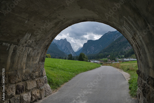 Blick durch eine gemauerte Unterführung auf einen Radweg der in Richtung eines Ortes mit Bergpanorama im Hintergrund läuft.