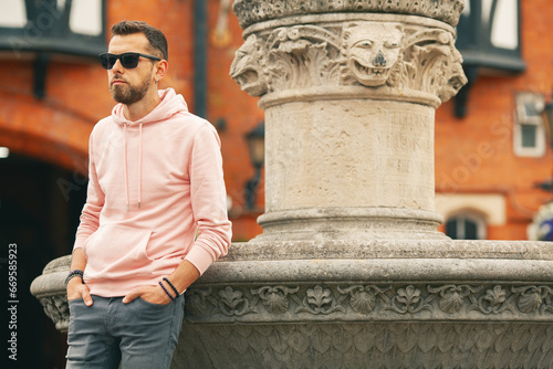 Travel concept. Young bearded romantic man wearing pink trendy hoodie, gray jeans posing over brick house with flowers on backgound. Old classical fontain. Outdoor shot