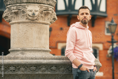 Welcome concept. Young smiling romantic man wearing pink trendy hoodie posing over brick house with flowers on backgound. Old classical fontain. Outdoor shot
