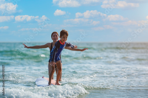 Happy little child boy learning to play surfboard from his mother with having fun and passion in sea on summer vacation at tropical beach.