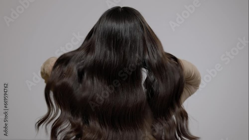Charming brunette woman shaking her long wavy hair on a light gray background. Back view.