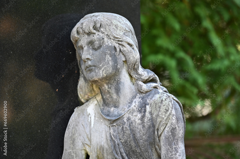 Partial view to a very weathered sandstone sculpture of a female angel face with a gentle look, reflecting on a  tombstone on a cemetery in Weimar-Germany. 