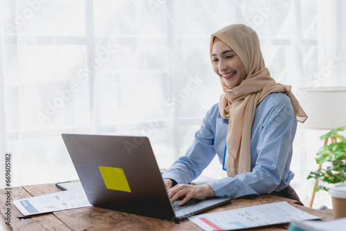 Muslim young woman sit at her desk with a laptop and document in the office with a bright smile and cheerfulness at work.