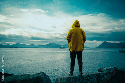 Guy standing on the rock and admiring a beautiful scenery of Norway fjord and mountains. Rainy weather and warm clothing.
