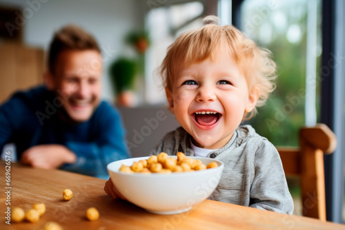 Happy toddler child boy eating a small bowl full of food. Parent is looking after him on the background