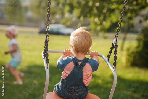 Baby kid in a denim jumpsuit swings on a swing in the summer in a public park, a place for text. The concept of a happy childhood. Active outdoors leisure for child.