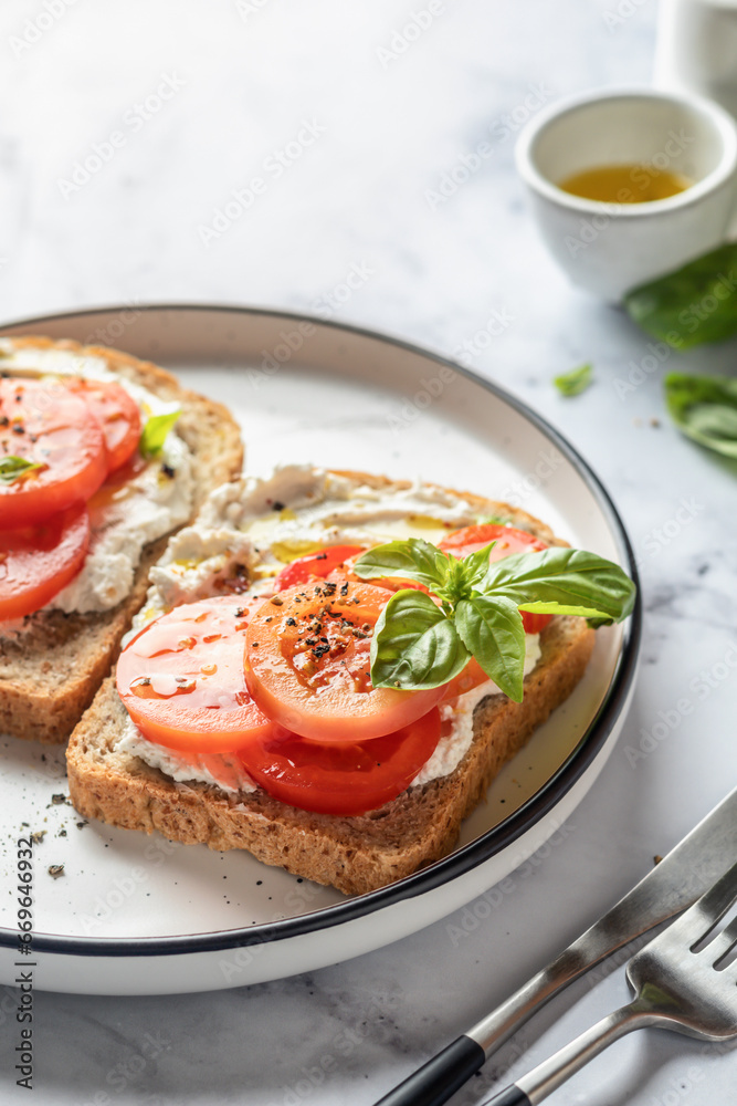 Sandwiches or toasts with tomatoes, cream cheese, olive oil and basil on a plate on white marble background. Traditional italian mediterranean food
