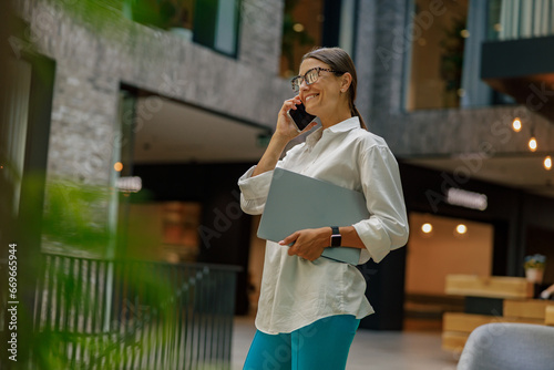 Business woman talking phone with client while walking in office hall with laptop