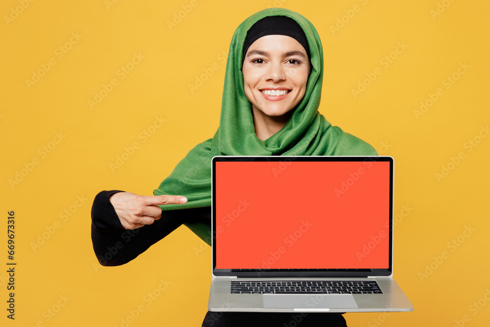 Naklejka premium Young asian muslim IT woman in green hijab abaya black clothes work use point on blank screen laptop pc computer isolated on plain yellow background People uae middle eastern islam religious concept