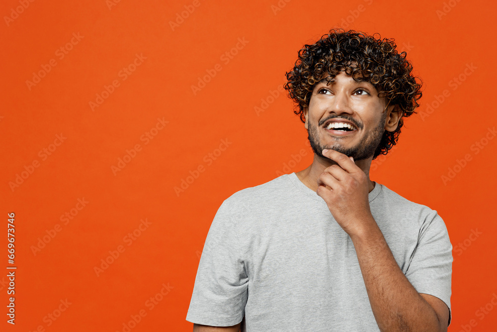 Young smiling happy Indian man he wearing t-shirt casual clothes put hand prop up on chin, lost in thought and conjectures isolated on orange red color background studio portrait. Lifestyle concept. Young smiling happy Indian man he wearing t-shirt casual clothes put hand prop up on chin, lost in thought and conjectures isolated on orange red color background studio portrait. Lifestyle concept.