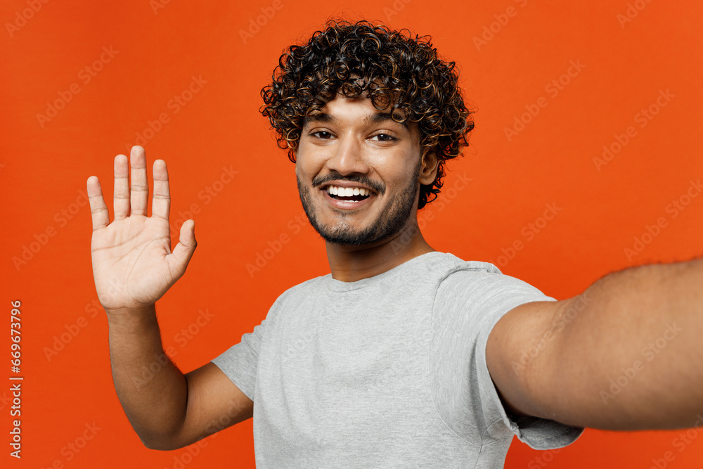 Close up young smiling cheerful happy Indian man wears t-shirt casual clothes doing selfie shot pov on mobile cell phone waving hand isolated on orange red color background studio. Lifestyle concept. Close up young smiling cheerful happy Indian man wears t-shirt casual clothes doing selfie shot pov on mobile cell phone waving hand isolated on orange red color background studio. Lifestyle concept.