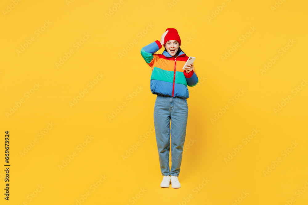 Full body young astonished cool happy woman she wearing padded windbreaker jacket red hat casual clothes hold head use mobile cell phone isolated on plain yellow background studio. Lifestyle concept. Full body young astonished cool happy woman she wearing padded windbreaker jacket red hat casual clothes hold head use mobile cell phone isolated on plain yellow background studio. Lifestyle concept.