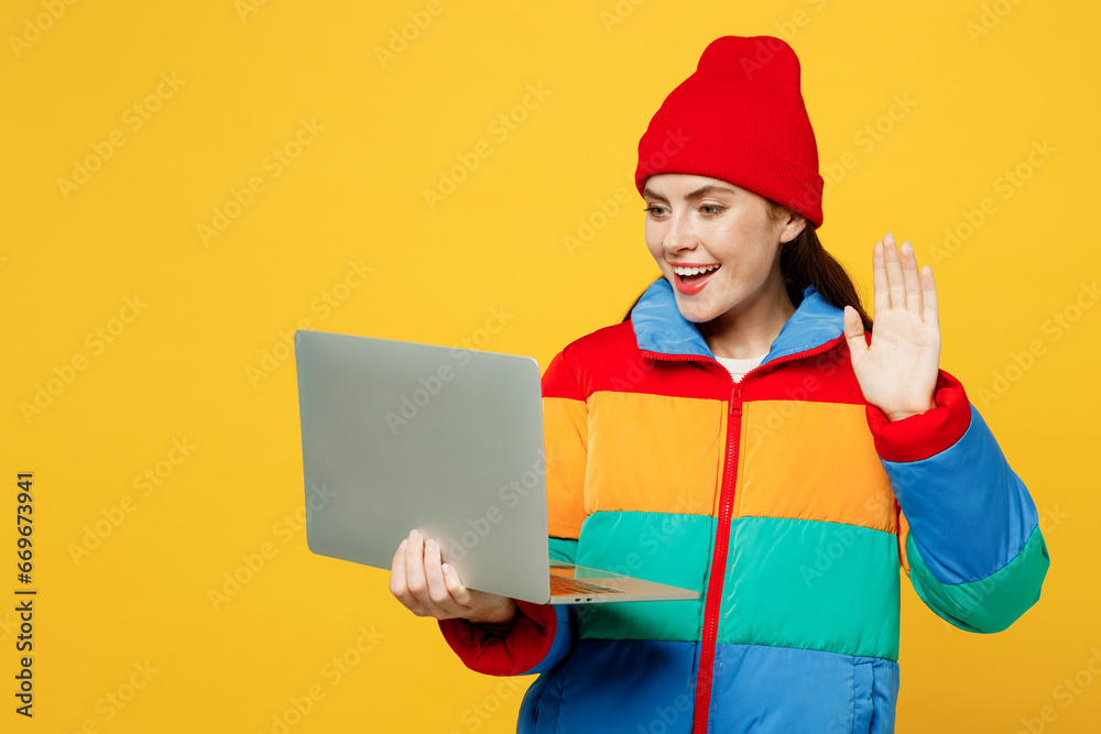 Young smiling fun IT woman she wearing padded windbreaker jacket red hat casual clothes hold use work on laptop pc computer waving hand isolated on plain yellow background studio. Lifestyle concept. Young smiling fun IT woman she wearing padded windbreaker jacket red hat casual clothes hold use work on laptop pc computer waving hand isolated on plain yellow background studio. Lifestyle concept.