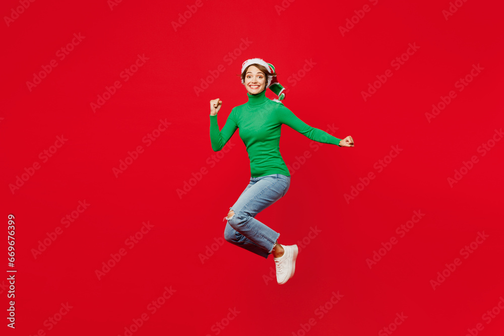 Full body young woman wear green turtleneck Santa hat posing jump high point index finger aside on mock up isolated on plain red background. Happy New Year 2024 celebration Christmas holiday concept.