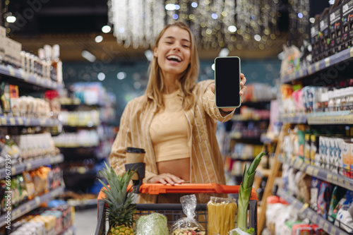 Young customer woman wear casual clothes showing blank screen mobile cell phone shopping at supermaket store grocery shop buy with trolley cart choose products in hypermarket. Purchasing food concept.