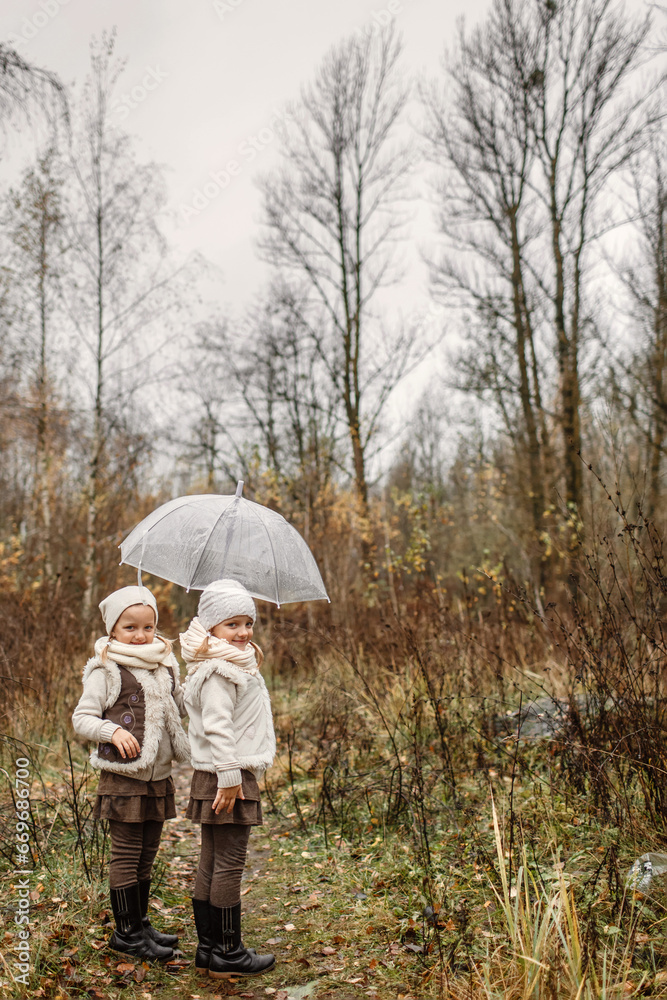 Portrait of young twin girls standing under an umbrella in the park, looking at the camera. Portrait of young twin girls standing under an umbrella in the park, looking at the camera.