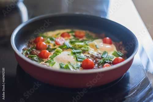 cook scrambled eggs with onions tomatoes and meat in a frying pan on an electric stove