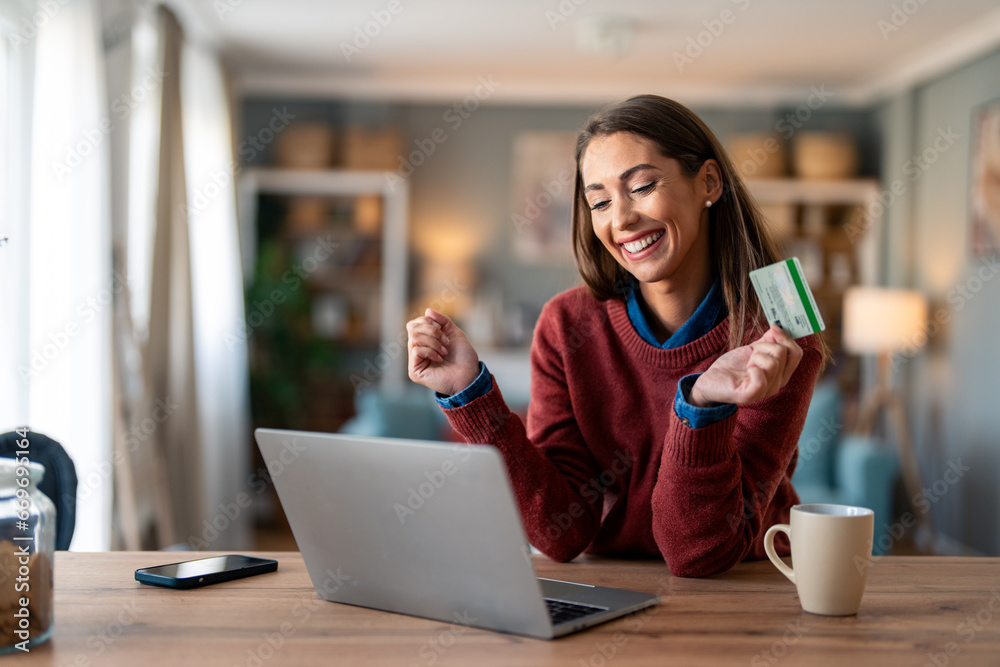 Happy young female shopper holding credit card, using laptop doing online banking transaction while sitting at desk at home. - obrazy, fototapety, plakaty 