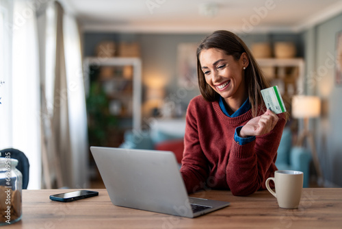 Canvas Print Smiling young adult woman holding credit card while shopping online over laptop computer