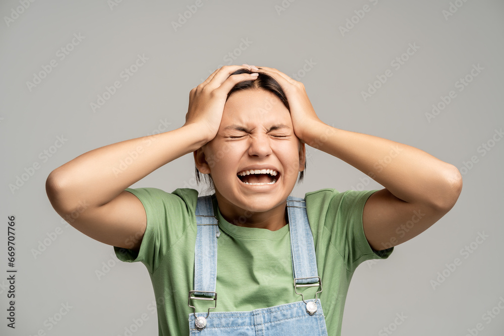 Sad desperated teen girl shouting holding head feeling intensive negative emotions on gray background. Teenager with closed eyes go through heartbreaking grief loss death of close people. Teen crisis. Sad desperated teen girl shouting holding head feeling intensive negative emotions on gray background. Teenager with closed eyes go through heartbreaking grief loss death of close people. Teen crisis.