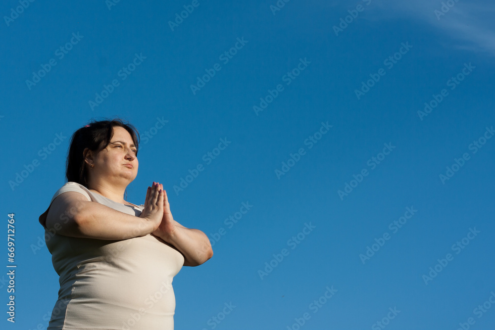 Portrait of middle-aged woman against blue sky. Calm woman holding hands in namaste mudra during meditation. Overweight woman practices yoga outdoors in summer. Mental health, healthy lifestyle Portrait of middle-aged woman against blue sky. Calm woman holding hands in namaste mudra during meditation. Overweight woman practices yoga outdoors in summer. Mental health, healthy lifestyle